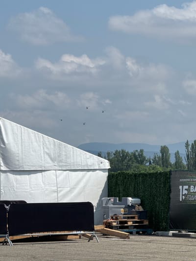 This image captures an outdoor scene, likely an event setup, under a partly cloudy sky. The perspective is from ground level, looking towards a horizon featuring distant mountains.
In the foreground, a large, white canvas tent dominates the lower-right portion, with part of its peak and side visible. To its left, a tall, black rectangular structure covered in fabric, possibly a speaker tower or stage element, stands on a paved surface, secured by metal scaffolding.
The mid-ground features a dark green, artificial hedge acting as a barrier. Behind it, a black banner displays Cyrillic text, partially reading "ИЗЛОЖЕНИЕ ЗА ЗА" (Exhibition for) and "15 BA ГОДИ" (15th Years), suggesting a 15th-anniversary exhibition or event. Below the banner and behind the hedge, various equipment is visible, including a gray utility unit on wheels, likely a generator, and stacks of wooden pallets, along with some loose wooden planks. The ground in the foreground is paved, suggesting an event space or parking area.
In the background, hazy blue-gray mountains rise, partially obscured by tall green trees. The sky above is a pale blue, dotted with soft, white and light-gray cumulus clouds. Against this sky, at least four small, dark, kite-like objects, resembling distant paragliders or drones, are visible, seemingly in flight.
The scene takes place during daylight hours, with diffused lighting from the cloudy sky. The overall atmosphere appears calm and organized, possibly during the setup or an ongoing, casual outdoor event in Madzherito, Bulgaria. There are no people clearly visible.