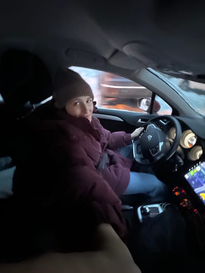 The image depicts the interior of a vehicle in Lesnoy Gorodok, Russia, with a female driver in motion, looking back at the camera. She is wearing a brown knit beanie and a dark maroon puffy winter jacket over light blue pants. Her right hand is positioned on the black, multi-function steering wheel, which features a Citroën logo. To the right of the steering wheel, circular instrument gauges are visible, and below them, an illuminated infotainment screen displays a navigation map interface with green, yellow, and red lines. The exterior background, visible through the windshield, is horizontally blurred, indicating vehicle movement. Snow is present on the ground outside, suggesting winter conditions and low ambient light. A hand, belonging to the front passenger (presumed photographer), is visible in the lower left foreground.