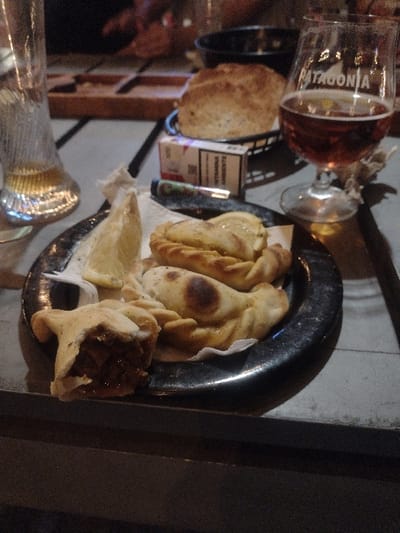 A plate of empanadas rests on a black metal surface alongside a partially visible beer glass displaying "Patagonia" lettering and a smaller glass. A piece of bread and a cigarette packet are nearby. The empanadas are golden brown, some slightly burned, with one visibly torn open to reveal its filling. A slice of lemon sits among the pastries on a white napkin.