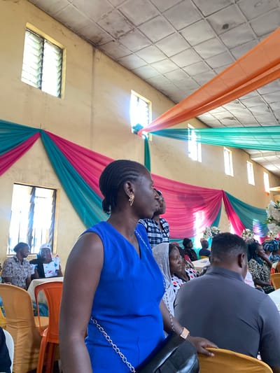 In Turu, Nigeria, an indoor event is taking place in a hall decorated with draped fabric in orange, teal, and pink. Several individuals are seated, some facing away from the camera and others looking towards the left. The main subject, a Black woman wearing a blue sleeveless dress and a gold star-shaped earring, is prominently positioned on the right side of the frame, her profile facing left. She is holding a black handbag. To her left, a Black man in a grey shirt is seated, also facing away from the camera. In the background, more attendees are visible, seated at tables. The hall has a tiled ceiling and windows with louvres, indicating a functional, rather than luxurious, setting. The lighting appears to be natural light filtering through the windows, supplemented by interior lighting that creates a warm ambiance.