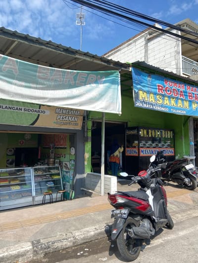 The scene is a street in Tulangan, Indonesia, with a bakery on the left side and a restaurant on the right. The bakery is called "A Bakery" and has a green awning and a large window display filled with pastries. The restaurant is called "Rumah Makan Roda Baru" and is decorated with a bright blue sign with red lettering. The restaurant has a sign on the door that reads "RM. Roda Baru" and another sign that reads "Padang Masakan Padang" in a more rustic style. A woman stands in front of the restaurant in front of a dark door, wearing a white and green striped shirt with a yellow cardigan and brown pants. She is standing near a table with a yellow and red tablecloth. There is a black motorcycle parked in front of the restaurant. There is a second black motorcycle parked on the sidewalk, partially obscured by a sign that reads "Padang Masakan Padang". The motorcycles are both modern models with bright headlights and red accents. The scene is bright and sunny.  The sidewalk in front of the businesses has a light brown, textured concrete surface, and the street is asphalt. The scene is framed by a white, brick building in the background, partially obscured by a blue sky and white clouds.  There are several electrical wires running along the top of the buildings.  There is a tall antenna on top of the brick building.  The ground and sidewalk are dry and there is no evidence of rain.  The scene appears to be captured on a relatively clear day. The coordinates provided are -7.482 latitude and 112.648 longitude.