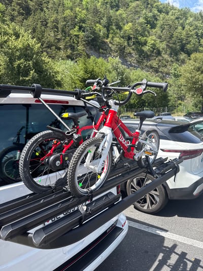 The image shows a close-up, eye-level view of a black bicycle rack mounted on the rear of a white vehicle, likely in a parking lot. Two children's bicycles are secured to the rack. The bicycle on the left is red and white, with the brand name "woom" visible on the frame. The bicycle on the right is white, and its front wheel is partially obscured. Both bicycles have knobby tires suitable for off-road use. The rack itself is made of black metal with individual channels for securing the bike wheels and straps to hold them in place. In the background, a white SUV with a sleek design is parked to the right, and a darker SUV is parked behind it. The environment appears to be outdoors, with lush green trees and a rocky hillside visible in the distance, suggesting a location with natural scenery, possibly on the outskirts of a town. The lighting indicates a sunny day. The scene is forensic in its detail, capturing the objects and their arrangement without any interpretation or speculation beyond what is visually present.