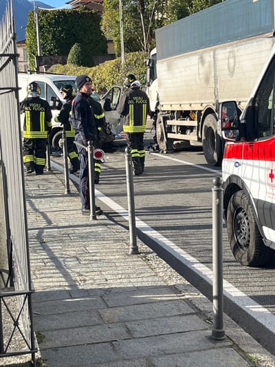 The scene depicts the aftermath of a multi-vehicle collision on a paved road in Griante, Italy, during daylight hours under a clear sky.
Four individuals, identified as "Vigili del Fuoco" (Italian Fire Brigade) personnel by their reflective yellow and black uniforms, are present near a severely damaged white van positioned on the left side of the road. The van's front end is heavily crushed, and debris is scattered on the asphalt.
Adjacent to the van, a large white and grey truck with a blue tarp covering its cargo bed is partially visible on the right side of the road. Its front end is also involved in the incident.
In the foreground on the far right, the front section of a white ambulance belonging to the "Croce Rossa Italiana" (Italian Red Cross) is visible, indicated by its red cross insignia.
A fifth individual, wearing a dark uniform and a dark beanie, stands on the paved sidewalk in the foreground, holding a circular red and white traffic control sign. Multiple dark bollards line the edge of the sidewalk, separating it from the roadway. A metal fence or railing is partially visible in the extreme left foreground.
The background features green foliage, residential buildings with terracotta roofs, and distant mountains. Long shadows cast across the sidewalk suggest a low to moderate sun angle.