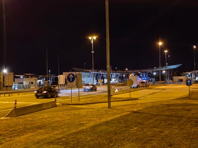 This image depicts a nighttime scene at a border crossing, likely Obrežje, Slovenia. The setting is an outdoor, paved area illuminated by streetlights and overhead lighting from the border facilities. The sky is dark, indicating it is nighttime.

The primary subjects are vehicles moving through the border crossing. Two cars are visible on the road, one in motion and blurred due to the long exposure, and another slightly further back. A white truck with flashing blue lights, possibly an emergency or service vehicle, is parked to the right of the road, alongside a smaller yellow vehicle.

The border crossing infrastructure includes booths or checkpoints, with modern, covered structures and numerous lights. Several signs are visible, including a blue circular sign with a white upward arrow, indicating traffic direction. Below this sign, there is text which reads "razen za vozila DARS in nadzorne službe".

The overall atmosphere is functional and somewhat sterile, characteristic of a border crossing at night. The lighting casts a warm, yellowish glow on the ground and vehicles. The grass in the foreground is illuminated by this light, creating a contrast with the dark sky. There are no people clearly visible in the image. The activity taking place is vehicular traffic processing through the border.