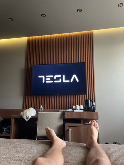 The image shows a hotel room interior in Otopeni, Romania, featuring a television displaying the Tesla logo, mounted on a brown wooden slat wall. To the right of the TV is a wooden cabinet with a coffee maker, cups, and other amenities. A plastic bag hangs from a shelf below the cabinet. A person is partially visible on a brown sofa, with bare feet and legs extending into the frame. The room has a light-colored wall with recessed lighting.