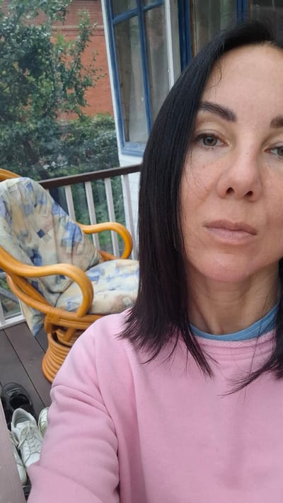 The image is a close-up selfie of a woman with dark hair, wearing a pink sweatshirt and a blue underlayer. She is on a porch or balcony with a railing visible. To her left, a wicker chair with a patterned cushion is present, along with a pair of black shoes and white sneakers on the wooden floor. In the background, green trees and a red brick building are visible through a blue-framed window. The overall setting suggests an outdoor, residential environment.