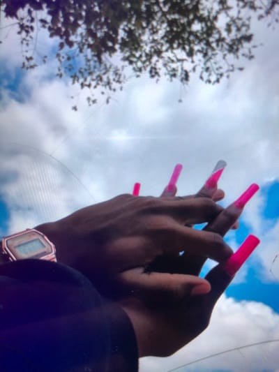 The image displays two intertwined hands with dark skin pigmentation, set against a bright blue sky with scattered white cumulus clouds. In the upper portion of the frame, blurred dark green tree leaves and branches are visible. The hand positioned on the left side of the image wears a rose gold-toned digital wristwatch on its wrist. The hand positioned centrally and to the right features elongated, squared, or coffin-shaped artificial fingernails, predominantly colored vibrant neon pink. One of these fingernails, possibly on the ring finger, includes a silver or white tip design. Diagonal and curved linear artifacts are present across the image, indicative of a photograph taken through a scratched or cracked surface. The scene is located outdoors in the city of Soweto, South Africa.