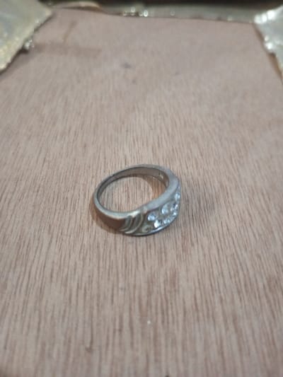 A silver-toned metallic ring is depicted resting on a light brown wooden surface. The ring features a broad top section; one half is adorned with multiple small, clear, faceted stones arranged in rows, while the other half displays an embossed swirling pattern. The metal exhibits minor surface wear. The wooden surface displays distinct vertical grain lines. The background is out of focus, revealing a blurred, lighter-toned area along the top edge of the frame. No individuals, actions, or interactions are visible. The location is Chandragiri, Nepal.