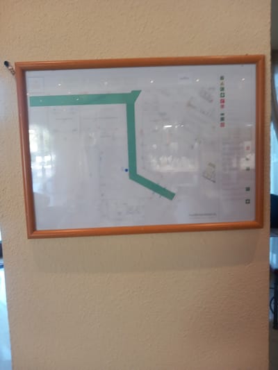 A framed diagram, likely a floor plan or emergency exit map, is mounted on a textured, light beige wall. The diagram features a prominent green pathway, indicating a route. To the right of the diagram, there is a vertical list of small colored symbols, including green squares, a red triangle, and red squares. The diagram is encased in a wooden frame with a warm, reddish-brown hue. The wall has a rough, stucco-like texture. The upper left corner of the frame is slightly disturbed by a black object, possibly a tack or pin, with a small silver component. Light reflections are visible on the surface of the diagram, suggesting it is under glass or a similar protective material. The overall scene appears to be indoors, possibly in a public building or establishment. The location is identified as the city of Kos, Greece.