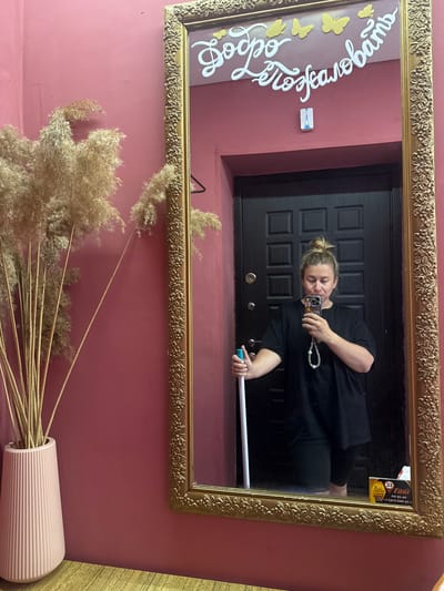 A woman is reflected in a large, ornate gold-framed mirror. She is standing in front of a dark, paneled door, holding a white mop handle in her left hand and a smartphone in her right hand, taking a selfie. She has blonde hair pulled up in a bun and is wearing a black t-shirt and dark shorts. A beaded strap hangs from her phone. The mirror is decorated with white lettering that reads "Добро Пожаловать" (Welcome) and several yellow butterfly cutouts. A small rectangular sticker depicting the Eiffel Tower is attached to the wall above the door. To the left of the mirror, a tall pink vase holds a large bunch of dried reeds. The wall behind the vase and mirror is painted a dusty rose color. A yellow and black taxi advertisement is visible on the lower right edge of the mirror. The scene appears to be indoors, possibly in an entryway or hallway. The city of Valday, Russia is mentioned as the location.