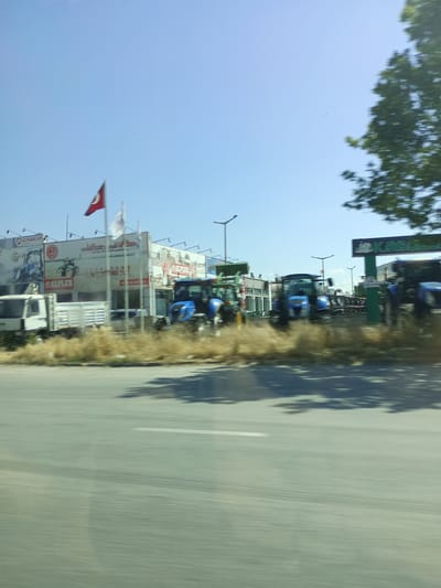 In Karatay, Turkey, a low-angle, blurred outdoor shot captures a clear sky with a few small birds. In the mid-ground, a building with several billboards displaying farm equipment and logos like "ALP ARSLAN" and "ALPİLER" stands. A white truck and several blue tractors are parked in front of the building. A Turkish flag is visible on a flagpole. In the background, more buildings, streetlights, and the blurry foliage of a large tree on the right side of the frame are present. The foreground consists of a road with a white line.