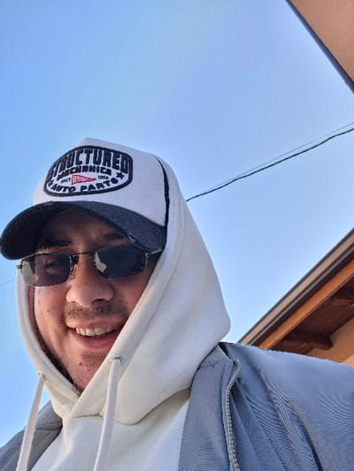 A male individual is depicted in an upward-angled outdoor photograph, positioned in Rakitovo, Bulgaria. He wears dark sunglasses, a white and black trucker hat featuring the text "STRUCTURED MECHANICS SINCE 1980 AUTO PARTS," a white hooded sweatshirt with the hood up, and a light grey zippered jacket. The individual is smiling, revealing teeth. The background primarily consists of a clear blue sky. A dark, thin utility wire or cable traverses horizontally across the middle-right section of the frame. The upper-right corner reveals a portion of a building's eaves, showing a light-colored fascia board and a darker underside.