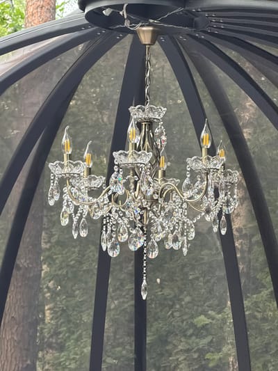 The image shows a crystal chandelier hanging inside a gazebo or similar outdoor structure with a black mesh roof and frame. The chandelier is ornate, with multiple crystal droplets and candle-shaped LED bulbs. The background is blurred, showing trees and greenery, suggesting an outdoor setting. There are no people or animals visible in the image. The location is described as Rakitnitsky Sel'ski Sovet, Belarus, though this specific detail cannot be verified from the image alone.