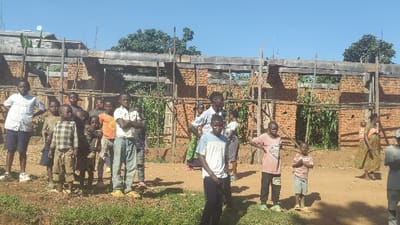 The image depicts a group of children standing in front of a partially constructed brick building with a wooden frame. There are 14 children present, all of African descent. Most are dressed in casual clothing. The children are standing in a group on a dirt path in front of the building, with some standing in front of the structure and others standing to the right and left. The building has a red brick exterior, a wooden frame, and some windows. The children are looking at the camera or at each other. The environment is rural, with green grass and trees in the background. The scene is located in Kayna, Democratic Republic of the Congo at the coordinates -0.608 latitude and 29.172 longitude.