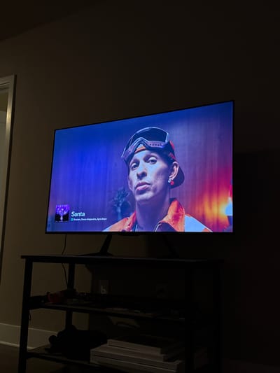 A TV screen displays a man wearing goggles and an orange jacket. The song "Santa" by Preston, Rauw Alejandro, and Ayra Starr is playing. To the right of the TV is a dark, multi-tiered shelf with various items on it, including stacks of white books or papers. The background is a dark wall.