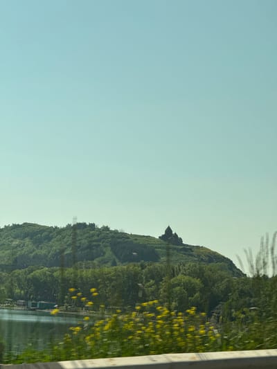 The image captures a scenic view of a lakeside landscape with a prominent monastery perched atop a lush green hill. The monastery, with its dark stone architecture and distinct dome, stands silhouetted against a clear, pale blue sky. The hillside is densely covered with trees, creating a vibrant green expanse. In the foreground, a calm body of water reflects the surrounding greenery. Along the shoreline, there are glimpses of small structures and what appear to be boats, suggesting a populated area. The overall scene is serene, with a sense of natural beauty and historical significance, likely located in or near Tsamakaberd, Armenia, given the architectural style and the characteristic landscape.