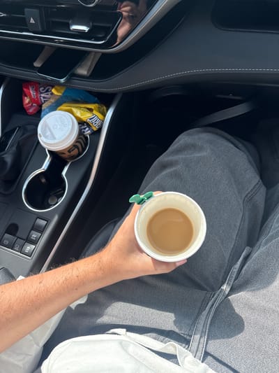 An overhead view inside a vehicle. A person is holding a disposable cup of coffee. There is a cup holder with another disposable coffee cup with a lid, a bag of Takis and a bag of RITZ crackers. A cell phone is in the side compartment. The car is in an unidentified location, not specifically San Rafael Tlanalapan, Mexico.