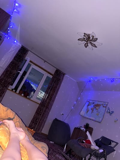 The image depicts an interior scene, likely a bedroom, illuminated by blue string lights. A window with patterned curtains is visible, showing a nighttime view with faint lights. A world map is mounted on the wall, along with decorative elements like a butterfly. Various pieces of furniture, including a beanbag chair and rocking chairs, are present. The overall ambiance is cozy and dimly lit, with the blue lights creating a distinct atmosphere. The image appears to have been taken in Leninogorsk, Russia, though specific details about the city are not discernible from the visual content alone.
