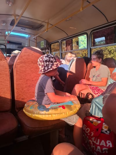 This image depicts the interior of a bus, likely in Russia, with several people visible. The overall scene suggests a relaxed or tired atmosphere, possibly during a journey.

In the foreground, a young child is seated, wearing a bucket hat with a panda pattern. The child is holding a yellow inflatable toy featuring a drawing of a crab. The child is also wearing a grey t-shirt and blue denim shorts.

Behind the child, a woman is asleep, leaning against the seat. She has light brown hair tied up and is wearing a light grey t-shirt with a graphic print and a red and white patterned skirt. Next to her, another person, partially visible, appears to be wearing blue clothing.

Further back, more passengers are seated, some appearing to be sleeping or resting. A red tote bag filled with items is visible, with some text on it.

The setting is the interior of a bus with brown upholstered seats and yellow metal poles for support. The windows show a glimpse of greenery and trees outside, suggesting it is daytime and possibly summer or early autumn. Sunlight is streaming into the bus, creating shadows.

Visible text on the red bag appears to be in Cyrillic script, potentially a brand name or slogan. A green sign with text is also visible on the wall of the bus. The overall mood is one of repose and perhaps travel fatigue.