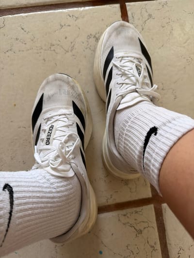 A pair of white Adidas Adizero running shoes are on a tiled surface. The shoes have black Adidas stripes and white laces. They are worn with white Nike socks. The tile floor appears worn, with visible cracks and a marbled texture. The photo is taken from a slightly overhead angle.