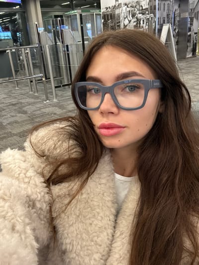 A woman with long brown hair and blue eyes, wearing gray-framed Gucci glasses, poses for a selfie. She's wrapped in a fluffy, cream-colored coat. The background features an airport terminal with security barriers, signs, and a grayscale image of a city landscape. The floor is a gray tile pattern. The picture is slightly rotated.