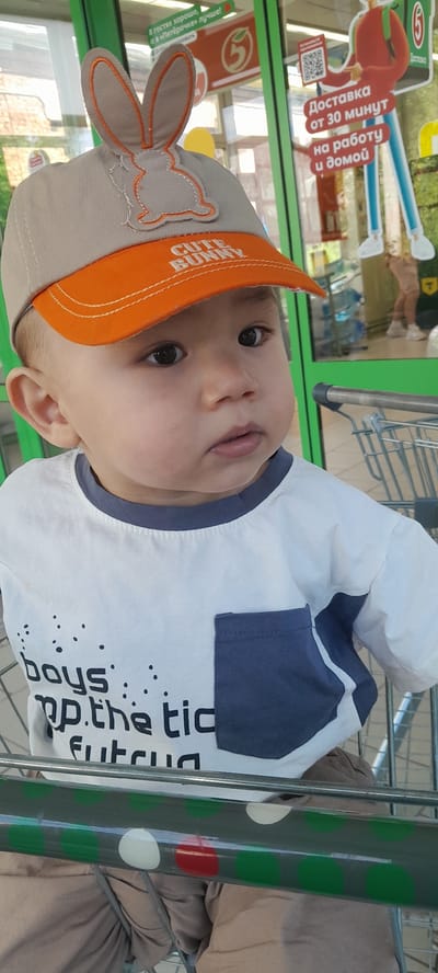 This is a close-up, vertically oriented, outdoor photo of a baby in a shopping cart. The baby is wearing a tan baseball cap with orange trim and bunny ears, and a white t-shirt with blue accents and black lettering. The baby has dark eyes and is looking directly at the camera with a neutral expression.

The background appears to be a retail environment, possibly a grocery store, with shelves and product displays visible through a glass partition. There is signage in Russian, with one prominent sign reading "Доставка от 30 минут на работу и домой" (Delivery from 30 minutes to work and home). Another sign says "В гостях хорошо, а в «Пятёрочке» лучше!" (It's good to visit, but it's better at Pyaterochka!). The overall lighting suggests it is daytime, with natural light contributing to the scene. The shopping cart itself is a metallic, wheeled cart, partially visible and framing the baby. The presence of a shopping cart suggests the baby is accompanied by an adult who is likely shopping. The scene is set in Pesyanka, Russia.