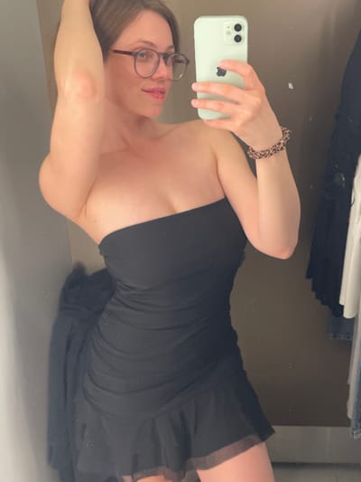A woman wearing glasses and a strapless black dress is taking a selfie in front of a mirror. She is holding a light green smartphone in her left hand. The background is a neutral-colored wall with some clothing items visible above her. The photo appears to be taken indoors. There is no information in the image to suggest the location is in Keszthely, Hungary.