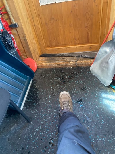The image captures an indoor utility area in La Salle-les-Alpes, France. The floor is dark grey with light blue speckles and appears wet. In the foreground, a human lower right leg is visible, clad in dark blue trousers and a brown lace-up hiking boot. A light brown wooden door, featuring vertical paneling and natural wood grain, is centered ahead, set within a matching wooden doorframe. A white document is affixed to the wall above the door. To the left, a dark blue, ribbed object, consistent with a sled or storage bin, rests on the floor. Behind this object, against the doorframe, a red circular item is present. Further left, a ski or snowboard with black bindings is propped against the wall, adjacent to a vertically leaning sledgehammer with a wooden handle. A red cord descends from the upper left. To the right of the door, a metal shovel with a D-handle and a red element is propped against the wall. Scattered water droplets are visible across the floor surface.