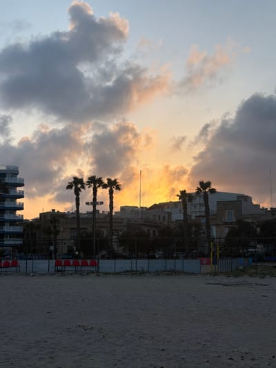 This image depicts a coastal scene in Birżebbuġa, Malta, at sunset. The foreground features a dark, unoccupied sandy beach. Immediately behind the beach, a low metal fence separates the sand from an area containing multiple rows of unoccupied red stadium-style seating. Further back, a series of five distinct, tall palm trees are silhouetted against the sky, standing amidst or in front of various urban structures. The midground also includes what appears to be a fenced-off sports court or similar ground-level facility.
In the background, a dense collection of buildings of varying heights and architectural styles line the horizon. These include a modern multi-story building on the far left with light blue accents and balconies, alongside more traditional structures. Several antennas and masts protrude from the rooftops of these buildings. The sky is dominated by a dramatic sunset, with a vibrant orange and yellow glow concentrated near the horizon, extending upwards and blending with dark grey and lighter blue clouds that cover the upper portion of the frame. No individuals are visible, and no actions or interactions are depicted within the scene.