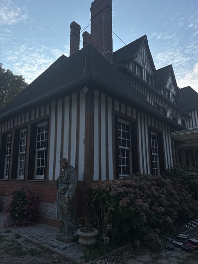 The image depicts a view of the side of a half-timbered building, likely a house or inn, in Gueutteville, France. The building features prominent dark wood beams against white infill, with several multi-paned windows visible. Two brick chimneys rise from the dark tiled roof. In the foreground, a weathered stone statue of a robed figure stands beside a large bush with purple flowers and a small potted plant. A cobblestone path leads around the base of the building. The sky above is partly cloudy.