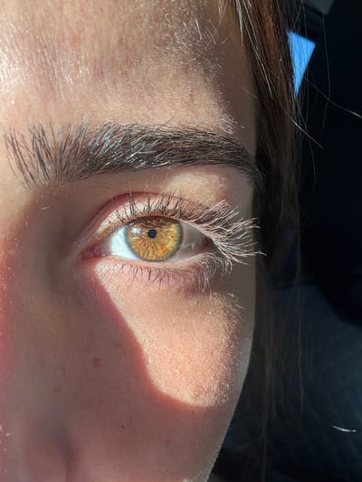 The image shows a close-up of a person's eye, tilted to the side. The eye has a hazel iris with a dark pupil. The upper and lower eyelids are visible, with eyelashes. The skin surrounding the eye is fair, with some shadows cast across the face. Some hair is visible at the top of the frame. There is not enough information to give the specific location in the city of Cidade da Maia, Portugal.