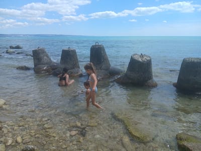In Osenovo, Bulgaria, on a sunny day, several concrete tetrapods protrude from the shallow, clear waters of the Black Sea. Three children are visible in the water near the tetrapods. The child closest to the foreground, a girl with brown hair tied in a ponytail, wears a light-colored bikini and walks through the water. To her left, a younger child, also in a light-colored bikini, is partially submerged in the water. Further to the left, another child with dark hair sits in the water, facing away from the camera. The seabed is visible and is covered in small stones and rocks. In the distance, the shoreline is visible with hints of vegetation, and the sky is a clear blue with scattered white clouds. The sea is calm and a bright turquoise color.