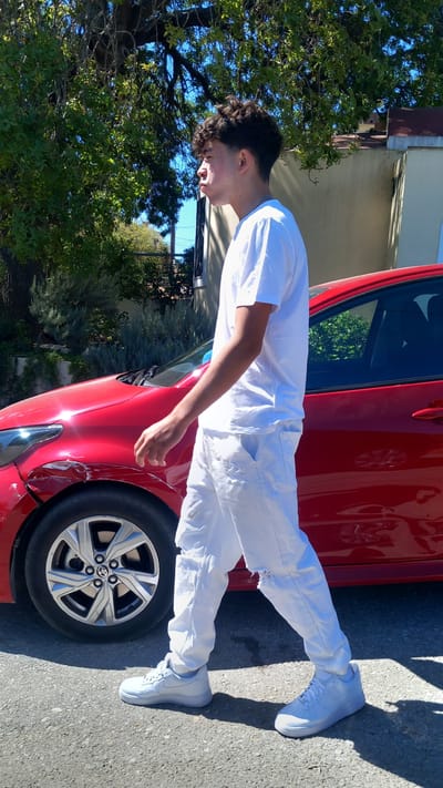 A young man with curly dark hair, wearing a white t-shirt and distressed white jeans, walks alongside a dented red car on a sunny day. The car, possibly a sedan, has visible damage to its front fender and bumper on the driver's side. The background shows green trees and a building with a terracotta roof, suggesting a residential or semi-urban area. The pavement appears to be asphalt. The man is looking towards the left of the frame, with his mouth slightly puffed out. The lighting indicates a bright, outdoor environment. The location is identified as Platani, Greece.