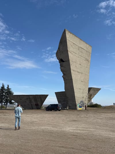 The image displays the "Monument to Liberators" located on Mount Kremenets in Izium, Kharkiv Oblast, Ukraine, under a clear blue sky with scattered white clouds. The primary subject is a large, weathered concrete monument composed of multiple angular, Brutalist-style slabs situated on unpaved light brown earth. The most prominent slab, centrally positioned on the right, is tall and angular, featuring visible horizontal striations from its construction and irregular, jagged damage along its left vertical edge. The base of this slab displays spray-painted graffiti: the word "МЕДОВА" (MEDOVA) above a blue and yellow Ukrainian flag. To the left, a lower, triangular concrete slab also bears faint graffiti, including "СЛАВА" (SLAVA) and "ВОЛЯ" (VOLYA). A dark-colored Mercedes-Benz van is parked facing left on the dirt ground between these two concrete elements. In the foreground, a male individual is captured from behind, walking away from the viewer towards the monument. He wears a light blue long-sleeved shirt, blue jeans, and brown boots. Two evergreen trees are visible on the far left. A faint, distant figure is discernible on the far right horizon, and the background shows a flat horizon line over what appears to be a distant landscape or city.
