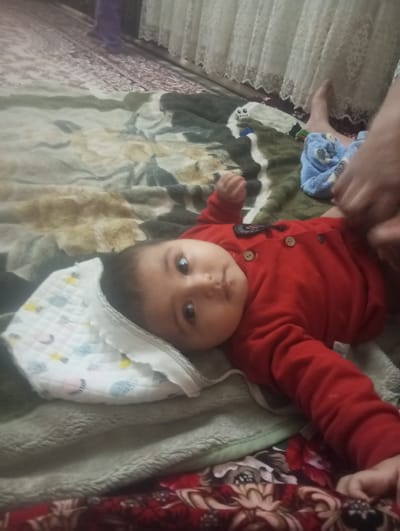 A baby wearing a red onesie is lying on a blanket with a patterned pillow under its head. A person is partially visible, near the baby's legs, likely changing or adjusting the baby's clothes. The setting is indoors, in a residence within the city of Galaosiyo, Uzbekistan. The environment includes a patterned rug and curtains in the background.