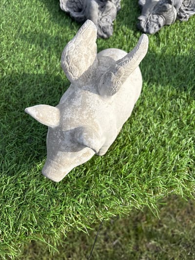 An aerial view shows a cement garden statue of a pig with wings on a patch of bright green artificial turf in Chirk, United Kingdom. Another, darker garden statue is partially visible at the left side.