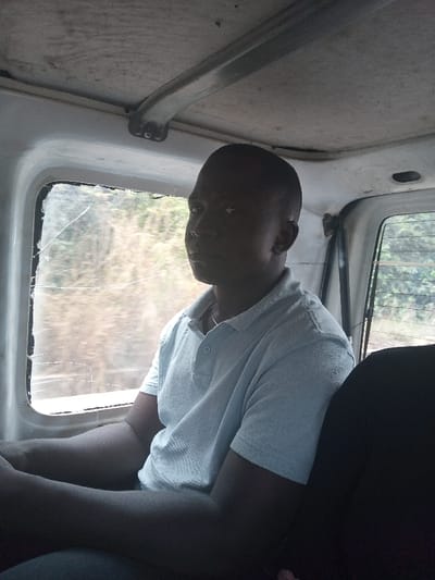 The image displays an adult male seated inside a vehicle, likely within or around Abor, Nigeria. The male, with short dark hair and a light complexion, wears a light blue short-sleeved polo shirt and a thin silver-colored chain around his neck. He is positioned facing slightly to the left of the viewer, his gaze directed forward or out the window. His left arm is visible, resting on his lap or the seat. The vehicle's interior has a light-colored ceiling with visible metal support structures and a white window frame. The side window adjacent to the male exhibits multiple distinct cracks. Through this window, an out-of-focus natural environment with green and brown foliage is visible. A dark-colored seatback is partially visible to the male's right. The scene is illuminated by ambient daylight, with the left side of the male's face appearing more brightly lit.