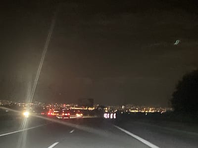 The image depicts a nighttime scene from a low-angle perspective, likely captured from a moving vehicle on a multi-lane road in the city of Yana, Bulgaria. The foreground is dominated by a dark asphalt road surface with distinct white lane markings, including a solid line on the right shoulder and a dashed line separating lanes to the left.

On the left side of the road, a powerful, overexposed light source, possibly an oncoming vehicle's headlight or a strong streetlamp, creates prominent vertical light streaks across the frame. Further ahead in the same direction, a line of red taillights from multiple vehicles is visible, receding into the blurred distance.

Along the right side of the road, a series of five white and red reflective road markers or barriers are arranged in a diagonal line. Beyond these markers, a dark, indistinct mass, suggestive of a tree line or dense foliage, occupies the far right of the frame.

The mid-ground and background feature the expansive, blurred lights of the city of Yana, extending across the horizon. These lights present as a bokeh effect, predominantly warm tones of yellow, orange, and white, with some scattered red points. Above the city lights, the sky is dark and appears slightly overcast. In the upper right quadrant of the image, a faint, light green, 'U'-shaped streak is visible against the dark sky. No individuals are discernibly present.
