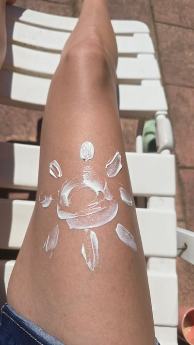 A close-up shot depicts a fair-skinned leg resting on a white plastic chair. The person has applied sunscreen to the leg, forming the shape of a sun. The leg is positioned towards the right, and part of a denim garment is visible at the bottom. The background features a patio-like surface, suggesting an outdoor setting. The presence of a sunscreen bottle implies someone is applying or has applied sunscreen for protection. No specific landmarks or architectural features are visible to locate the scene within Wilhelmshaven.