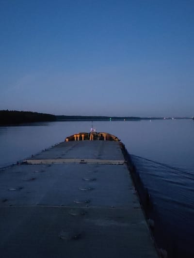 The image depicts a stern view from a large, flat-decked vessel navigating a wide body of water during twilight, near Yakunina Gora, Russia. The dark grey deck of the vessel extends from the foreground towards the center, featuring visible panel lines and several circular fixtures. Toward the far end of the deck, a cluster of illuminated yellow, cylindrical structures and a mast emitting a blue light are visible. A wake trails from the vessel's starboard side. A dense, dark forested shoreline forms the left bank of the waterway, receding into the distance. Scattered across the distant water, multiple lights are visible, including a prominent green light on the left and several fainter reddish-orange lights further to the right, indicative of navigation markers or distant shore features. The sky above is a deep, clear blue, transitioning subtly towards the horizon, consistent with low light conditions. No people are visible.