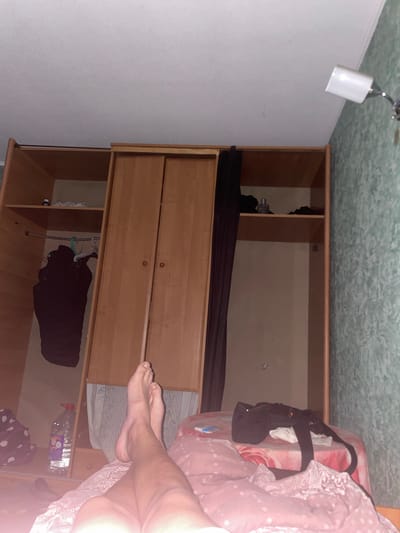 The image depicts an indoor scene, likely a bedroom, in Ivankovychi, Ukraine. The primary focus is a large wooden wardrobe with two doors, one of which is slightly ajar, revealing hanging clothes. To the left of the wardrobe, shelves are visible with various items, including a bottle of water and what appears to be clothing. On the right side of the wardrobe, the wall has a patterned wallpaper. In the foreground, a person's bare legs and feet are visible, positioned as if resting on a pink-covered surface, possibly a bed or sofa. A black bag is placed on the pink surface. A wall-mounted light fixture is visible on the ceiling in the upper right corner. The overall lighting suggests it is either nighttime or the room is dimly lit.