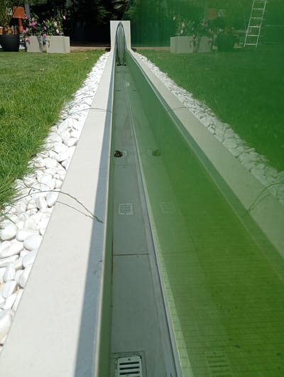 The image shows a long, narrow water feature with a light-colored concrete edge, bordered by white stones and green grass. The water in the feature has a green tint, possibly due to algae or a coloring agent. A glass or transparent divider is visible in the middle of the water feature, extending upwards. There are several square grates or drains visible along the bottom of the water feature. In the background, to the left, are potted plants with pink flowers and some dark foliage. Further back, a silver ladder is leaning against a structure, and there are trees and other greenery. The scene appears to be an outdoor area, possibly a garden or courtyard, in Nowa Wieś Szlachecka, Poland.