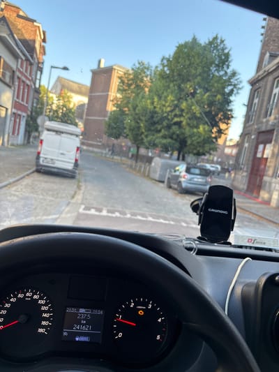 The image shows the interior of a vehicle, likely a van, looking out through the windshield. The view is slightly angled, with the steering wheel and dashboard visible in the foreground on the right. A black "GRUNDIG" phone mount is attached to the windshield.
Through the windshield, a street scene in a European town is visible, with buildings lining both sides of the road. The architecture suggests an older European city, consistent with the Liège, Belgium location context. There are several parked vehicles, including a white van in the middle distance. The sun appears to be shining, casting clear shadows and indicating it is daytime, with the dashboard clock showing 19:54.
The dashboard displays various readings. The speedometer shows the needle pointing around 30 km/h. The digital display shows the outside temperature as 29.5°C, a distance of 237.5 km, and an odometer reading of 241621. There is also a fuel gauge indicator. A small orange warning light is illuminated on the tachometer. The overall scene conveys a sense of normal daily activity within the vehicle, possibly driving through the town.