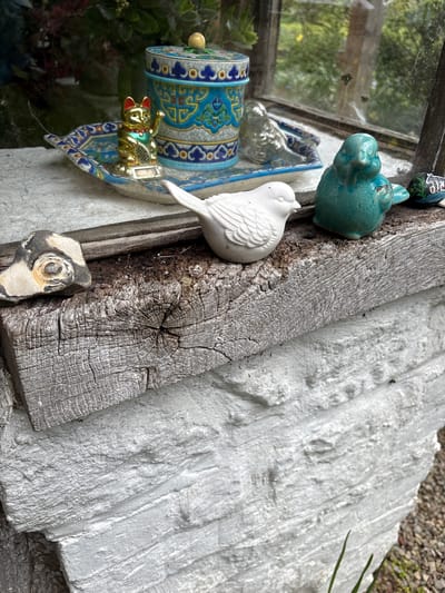 The image depicts a collection of small decorative objects arranged on or near what appears to be a windowsill and a rustic wooden plank. The objects include a blue ceramic bird, a white ceramic bird, a small decorative barrel-shaped object with intricate blue and gold designs, a golden cat figurine, and a piece of patterned rock. The background features a weathered white wall with a textured surface and a glimpse of greenery through the window. The location, Pulverbatch, United Kingdom, is based on the prompt but not directly observable in the image. There are no people depicted in the scene.