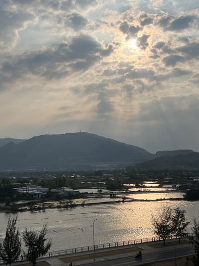 This wide-angle image depicts a scenic landscape, likely captured during sunrise or sunset, given the low sun position and the golden light illuminating the scene. The sky is a prominent feature, filled with dramatic, scattered clouds through which the sun actively breaks, casting bright, visible rays of light.
In the foreground and mid-ground, a tranquil body of water, possibly a river or bay, stretches across the frame. Its surface shimmers with intense golden reflections from the sun, creating a sparkling effect. On the immediate right bank of this water body, a paved road or pathway runs parallel, bordered by a black metal railing. Several dark, silhouetted trees, possibly coniferous, are visible along the path. A street light also stands near the road, and a dark shape that may be a parked motorcycle or scooter is discernible on the pavement near the railing.
Across the water, on the left bank, various buildings or houses are nestled at the base of a significant mountain range that forms the backdrop of the scene. The mountains appear hazy, suggesting atmospheric conditions or distance. A notable feature on one of the mountain peaks is a tall, slender structure, resembling a monument or statue, standing prominently against the sky.
The overall scene conveys a sense of peaceful natural beauty. Given the provided context, the location is the Nam Natural Heritage Area in Trang, Vietnam. No discernible text is visible within the image.