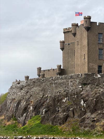The image depicts Dunvegan Castle, located in Dunvegan, United Kingdom. The castle is built on a rocky promontory overlooking a body of water, likely the sea. The castle itself is constructed of stone and features several towers and turrets. A flag is visible atop one of the towers. The surrounding environment consists of rocky cliffs and sparse vegetation. The sky is overcast. There is a hint of green in the bottom right corner, possibly vegetation. There are two figures standing on the outer castle wall.