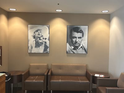 A medium shot captures a waiting room with two framed black and white portraits of actors displayed on a tan textured wall. The portrait on the left features a woman in a wide-brimmed hat and holding a bouquet of flowers, while the portrait on the right is a close-up of a man. Below the portraits are two brown leather sofas and two armchairs with textured brown upholstery. A round wooden side table with a magazine and a pen rests between the right sofa and an armchair. The room is lit by recessed ceiling lights, casting pools of light on the wall. The scene appears to be set in a commercial establishment or office, likely in Burbank, United States of America, given the overall aesthetic and the presence of office-like furnishings.