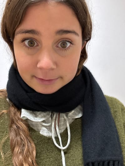 A head-and-shoulder shot features a young female subject looking directly into the camera. She has light brown hair, dark brown eyebrows, and wide, light brown eyes. Her mouth is closed with a slight upward curve at the corners. She wears an olive-green knitted sweater over a light-colored, possibly plaid or checked, collared shirt with a ruffled edge visible around the neckline. A loose white drawstring or tie hangs centrally from the shirt's collar. A black, finely woven scarf is wrapped around her neck, partially obscuring the lower portion of her chin and extending over her chest. The background is a plain, uniform white surface, indicative of an indoor setting. The specific location within Porto Salvo, Portugal, or any other city, cannot be determined from the image.