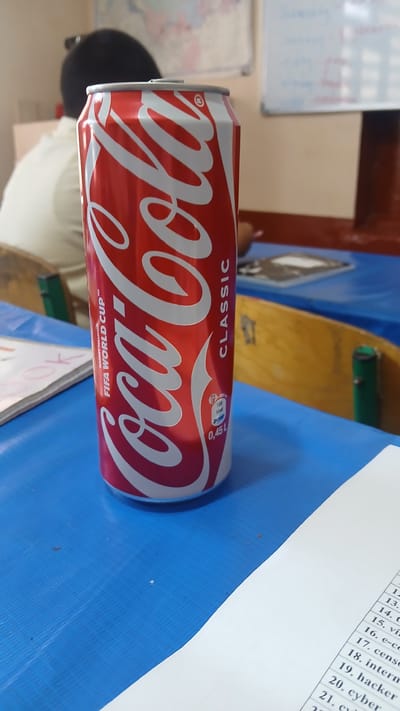 A close-up, eye-level, slightly low-angle shot captures a Coca-Cola Classic can standing upright on a vibrant blue, textured surface. The can, predominantly red with white lettering, features the iconic Coca-Cola logo and the text "FIFA WORLD CUP™". To the right of the can, the word "CLASSIC" is vertically oriented. Below "CLASSIC", there's a small circular graphic with nutritional information, showing "189 kcal" and "0,45 L". In the background, out of focus, a person with dark hair wearing a light-colored shirt is visible. Further in the background, a whiteboard with writing and a world map-like image on a wall are discernible. The scene suggests an indoor setting, possibly a classroom or meeting room, with multiple blue-covered tables and wooden chairs. The location is identified as Kattakurgan City, Uzbekistan.