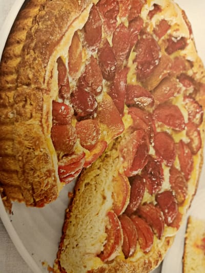 The image shows a close-up of a rustic fruit galette. The galette has a golden-brown, flaky crust that is folded over the fruit filling. The filling appears to be made of sliced plums, which are a deep reddish-purple color. The plums are arranged in a spiral pattern and are slightly caramelized from baking. A slice of the galette has been cut and partially removed, revealing the tender, golden crust on the side. The background is a plain white surface, likely a serving platter or table. There are no people or any identifiable landmarks visible in the image. The image does not provide any information about the location in Rumaithiya, Kuwait.